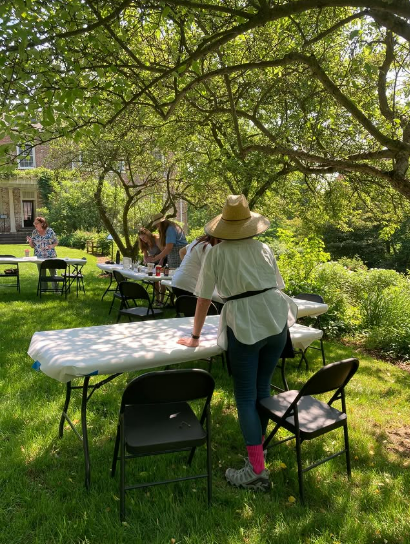 A woman in a sun hat stands at a table outside under a tree, facing several adults at other tables working on art projects.